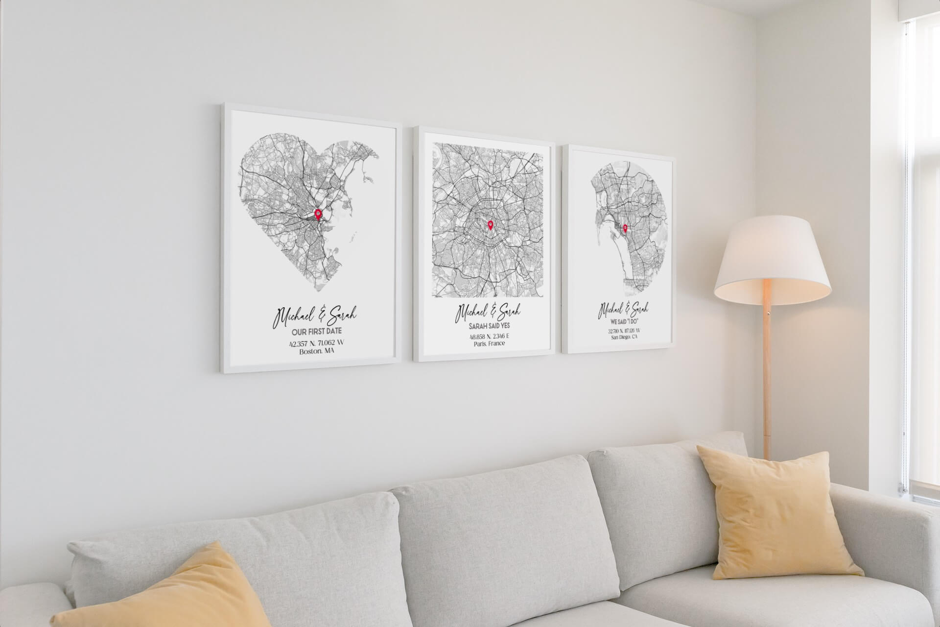 The image shows a living room with a light grey sofa and above the sofa are three framed posters with black-and-white maps and red location markers, depicting significant locations and dates for a couple named Michael and Sarah. The maps are titled "Our First Date," "Sarah Said Yes," and "We Said I Do," marking Boston, MA; Paris, France; and San Diego, CA, respectively. These personalized prints serve as unique gifts for anniversaries, capturing special moments in the couple's relationship.